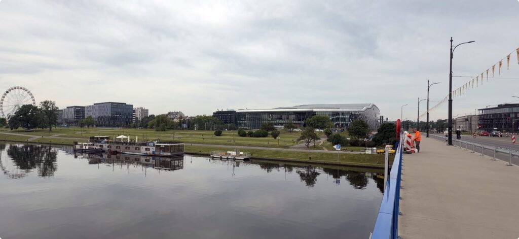 View from a bridge over a calm river or canal, with several moored houseboats reflected in the water. Across the water is a large grassy park bordered by modern office and conference buildings, including a distinctive curved glass-and-metal structure in the centre. A ferris wheel stands in the distance on the left, while a wide pedestrian walkway and road run alongside the bridge on the right under an overcast sky.